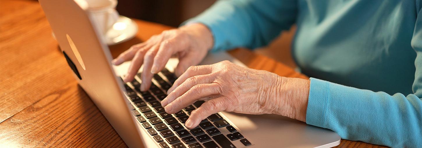 An elderly woman’s hands rest on the keyboard of a laptop.