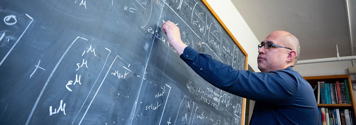 Man wearing glasses 和 blue shirt writes mathematical formula on blackboard.