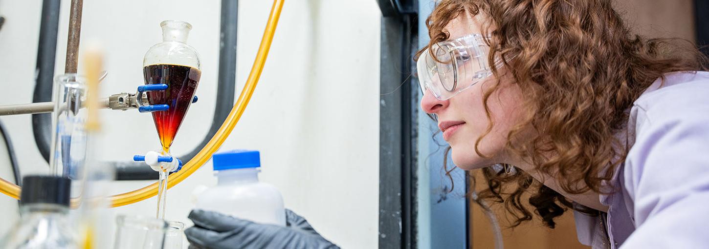 Woman in chemistry lab wearing safety goggles 和 lab coat examines a beaker.